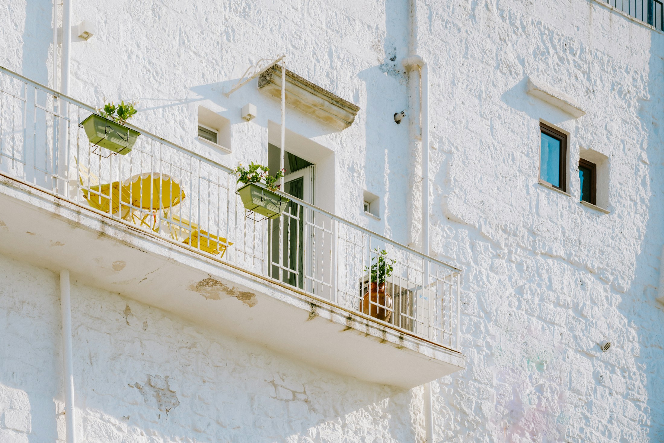 Biały kamienny balkon z kutą balustradą, żółtymi krzesłami i roślinami w doniczkach